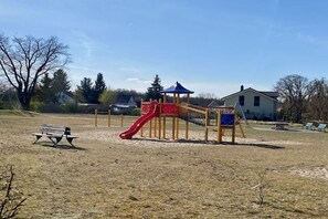 Ein Spielplatz mit Rutsche und Klettergerüsten, einer Bank sowie einem Haus im Hintergrund.