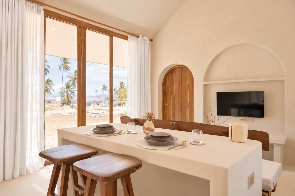 A dining area with a large window, a television mounted on the wall, and a view of palm trees outside.