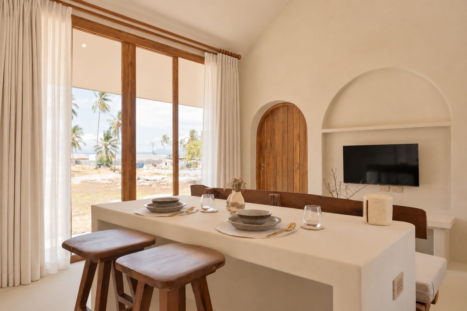 Un comedor con una mesa dispuesta para dos, un televisor montado en la pared y vistas a palmeras afuera.