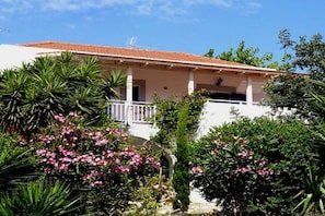 Un edificio blanco con techo de tejas rojas, un balcón y abundante vegetación, incluyendo flores rosas y palmeras.