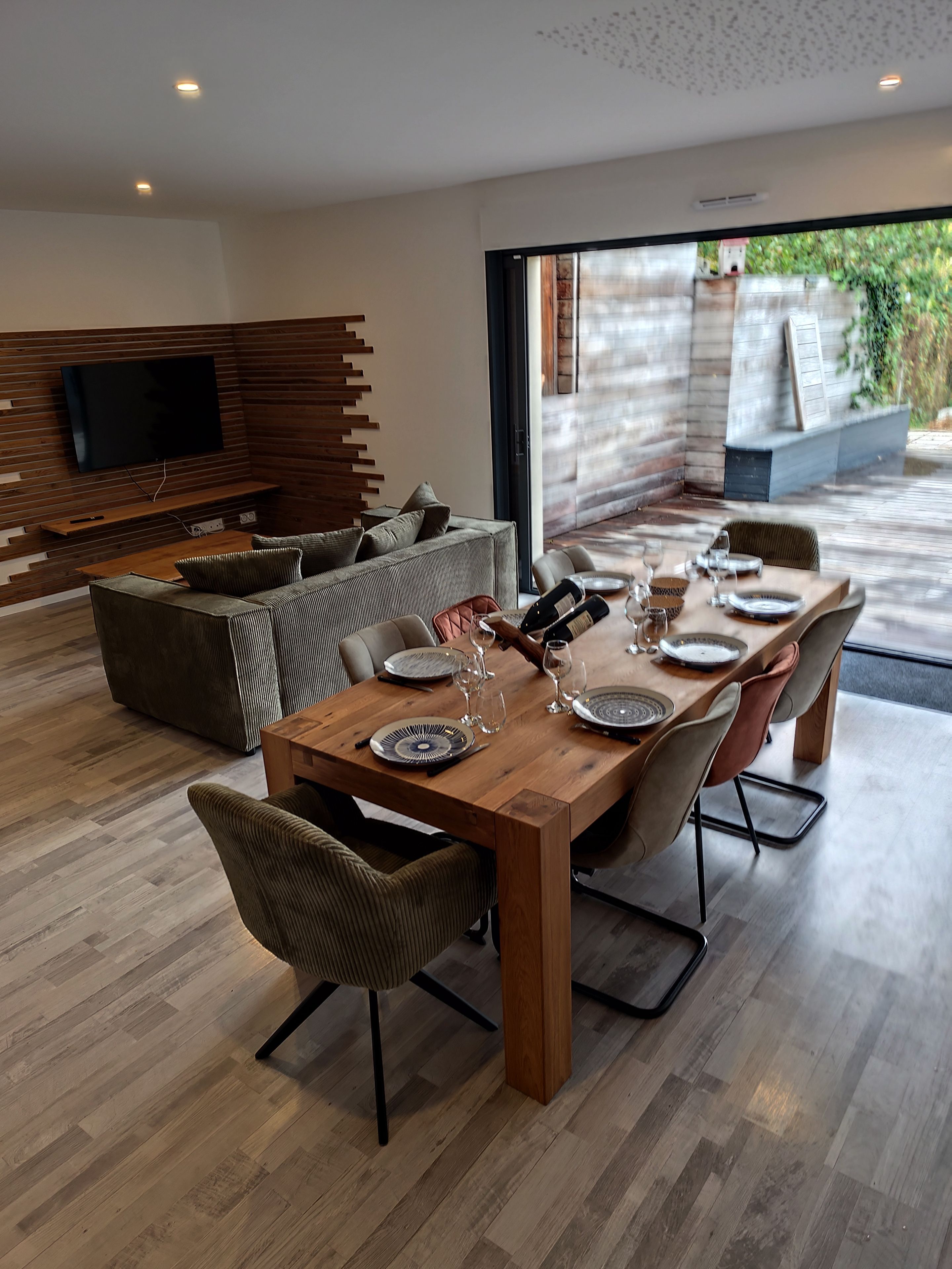 Un salon moderne avec une table à manger en bois, disposée pour accueillir six personnes, un téléviseur à écran plat fixé à un mur en bois, et une grande porte-fenêtre coulissante donnant sur un espace extérieur.