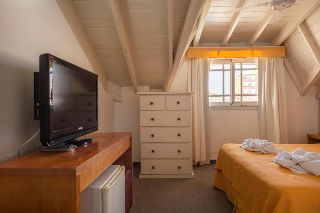 A bedroom with a television on a wooden desk, a white dresser, a bed with a yellow bedspread, and a window with curtains.