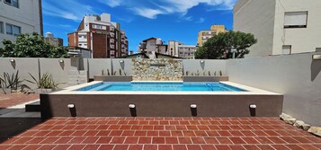 Una piscina rectangular con un muro de piedra de fondo, rodeada por un patio de ladrillo y edificios adyacentes.