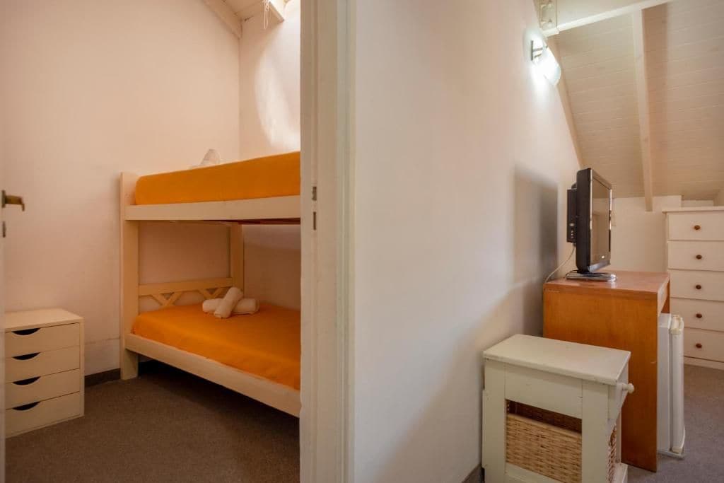 A bunk bed with a white and orange bedspread, a small white dresser with drawers, a television on a wooden stand, and a radiator.