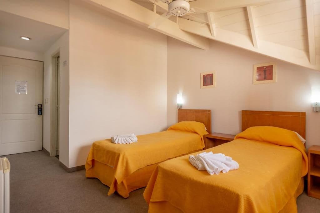 A hotel room with two beds, each with a white towel on the bed. There is a wooden headboard, a nightstand, and a wall-mounted light.