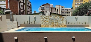A swimming pool with a stone wall and a water feature, surrounded by a low stone wall and plants. - Hotel Belmar (Villa Gesell)