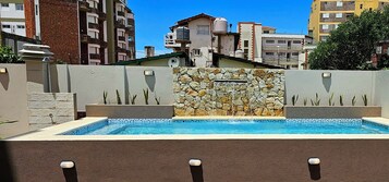 Una piscina con muro de piedra y fuente de agua, rodeada por un muro bajo de piedra y plantas.