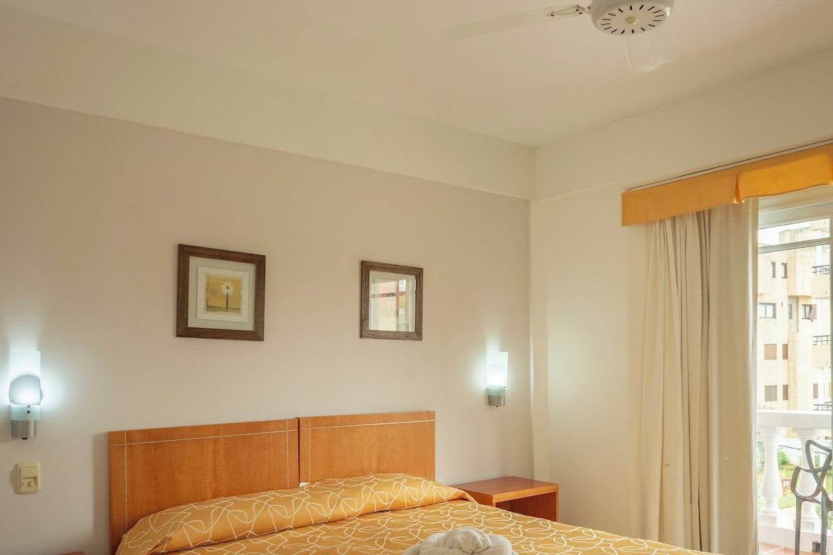 A hotel room with a large bed, a ceiling fan, two framed pictures on the wall, and a window with curtains.