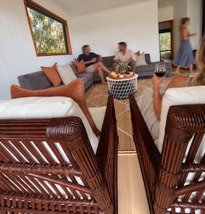 A modern living room with a sofa, chairs, a coffee table with a fruit and cheese board, and a large window with a view of trees. - The Lake House Coongulla - Lakefront Getaway for Couples (Coongulla)
