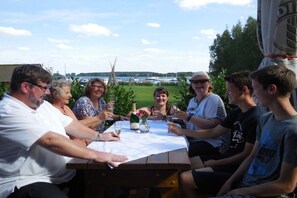 Un grupo de personas sentadas en una mesa al aire libre con mantel blanco, una botella de champán y un jarrón con flores.