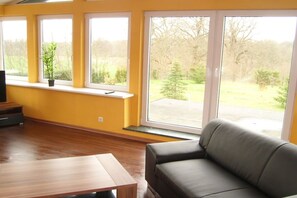 Ein Wohnzimmer mit einer Couch, einem Holztisch und einem Fernseher auf einem Schrank. Drei Fenster bieten Blick auf eine grüne Landschaft.