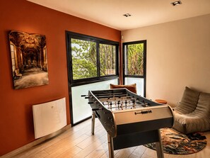 A room with a foosball table, a sofa, and a window with a view of trees. - Maison Romia by Sur la mer (Fréjus)