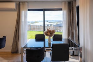 Ein moderner Essbereich mit einem Glastisch und schwarzen Stühlen, einer Vase mit Trockenblumen und Blick auf eine Wohngegend durch große Fenster.