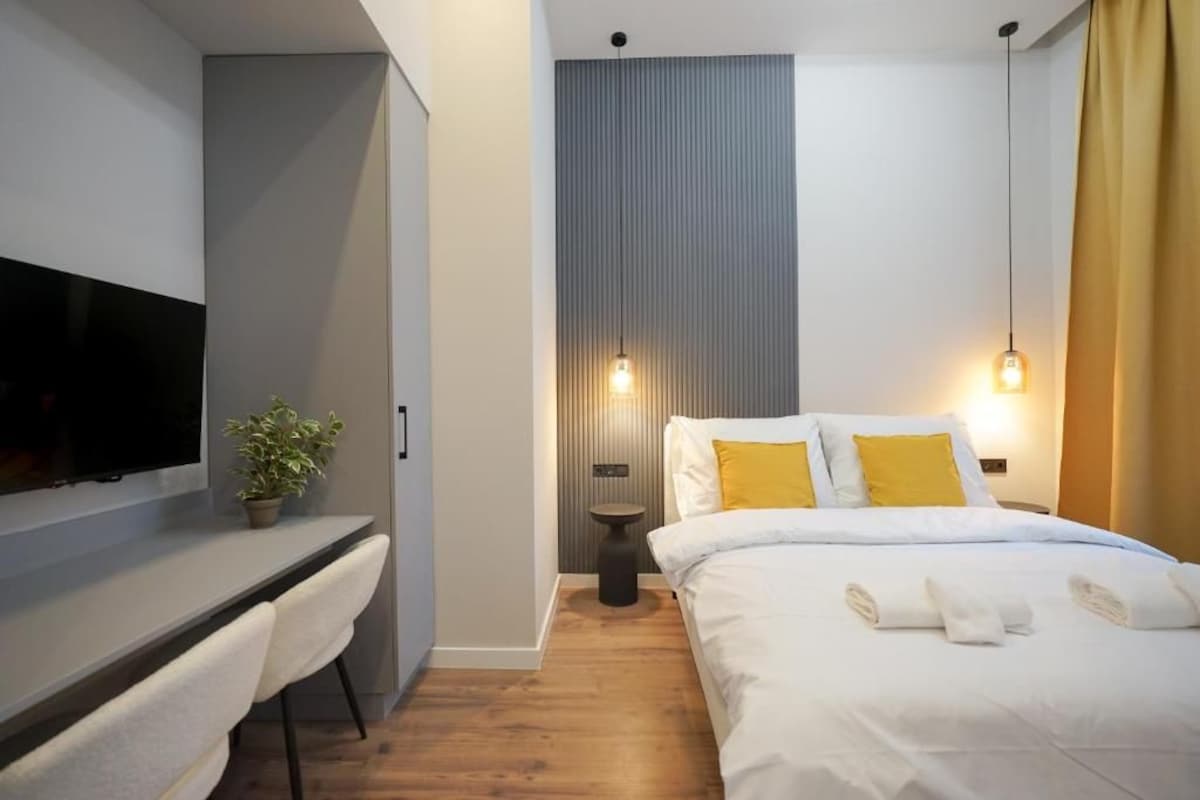 Een moderne hotelkamer met een flatscreen televisie, een bureau, een bed met witte beddengoed en gele kussens, een kamerplant en een wandlamp.