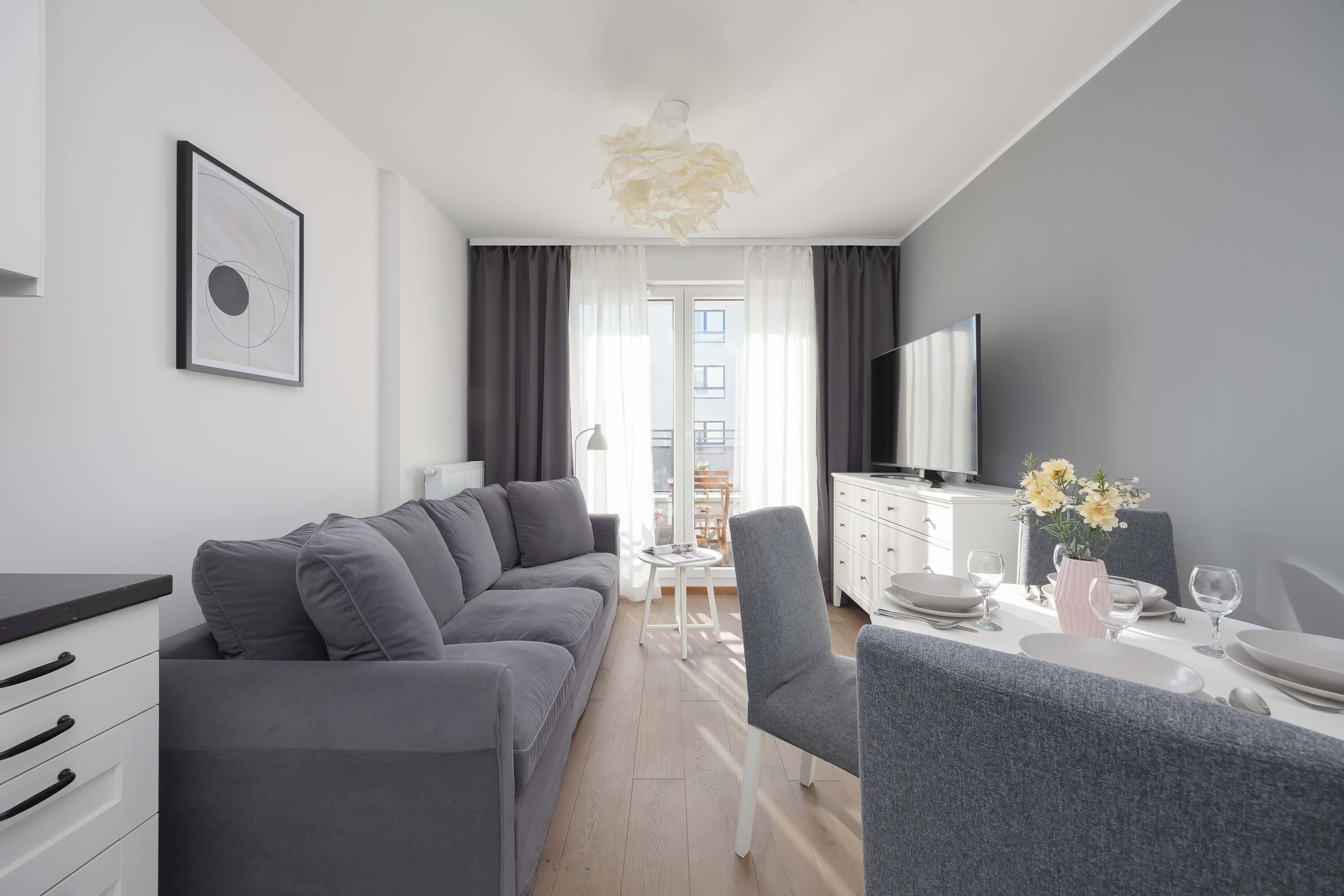 Ein modernes Wohnzimmer mit einem grauen Sofa, einem Esstisch für zwei Personen und einem wandmontierten Flachbildfernseher.