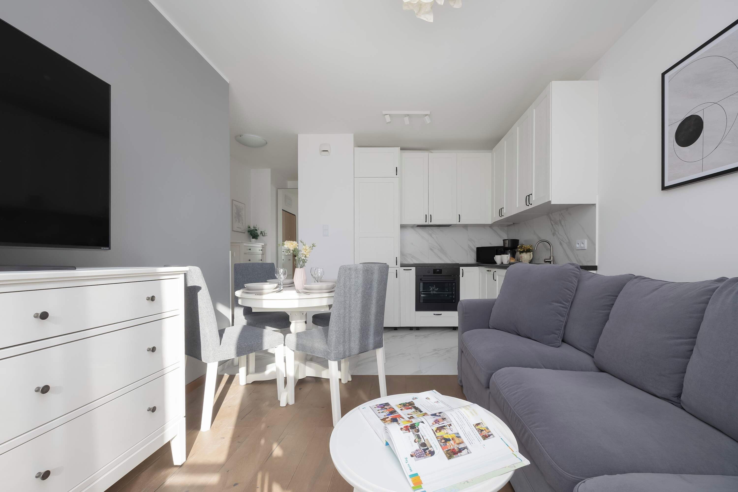 A modern living room with a grey sofa, a dining area with a white table and chairs, and a kitchen with white cabinets and a black oven.