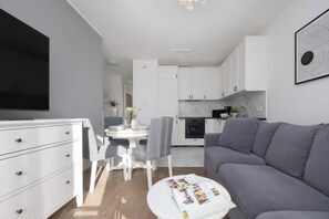 Ein modernes Wohnzimmer mit einem grauen Sofa, einem Essbereich mit weißem Tisch und Stühlen sowie einer Küche mit weißen Schränken und einem schwarzen Backofen.