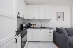 A modern kitchen with white cabinets, a black countertop, and a grey sofa in the adjacent living area. - Warsaw | Silnikowa 15 | Modern Apartment | Desk | Balcony (Warszawa)