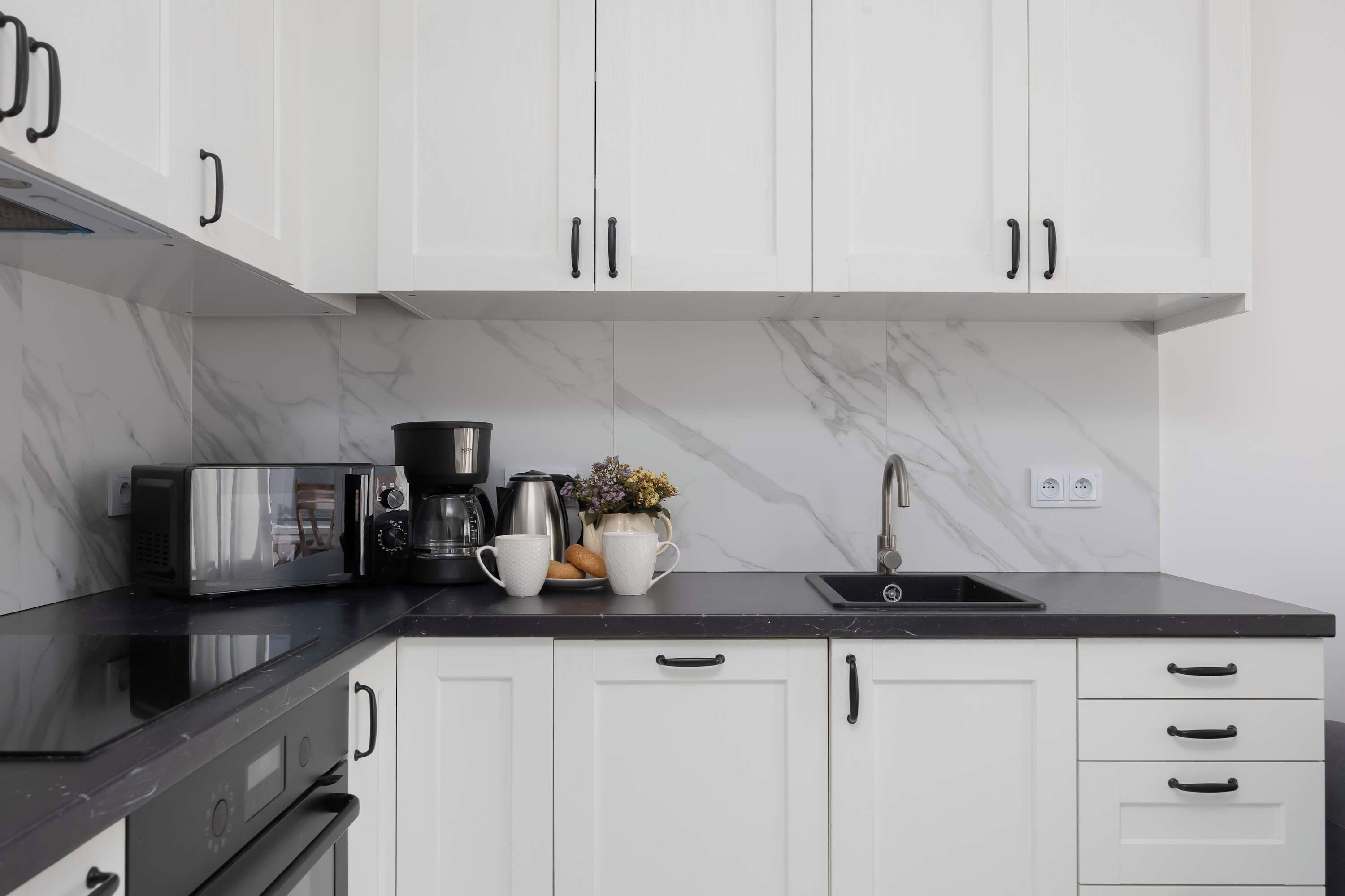 Una cocina moderna con armarios blancos, encimera negra, horno empotrado, microondas, cafetera y fregadero.