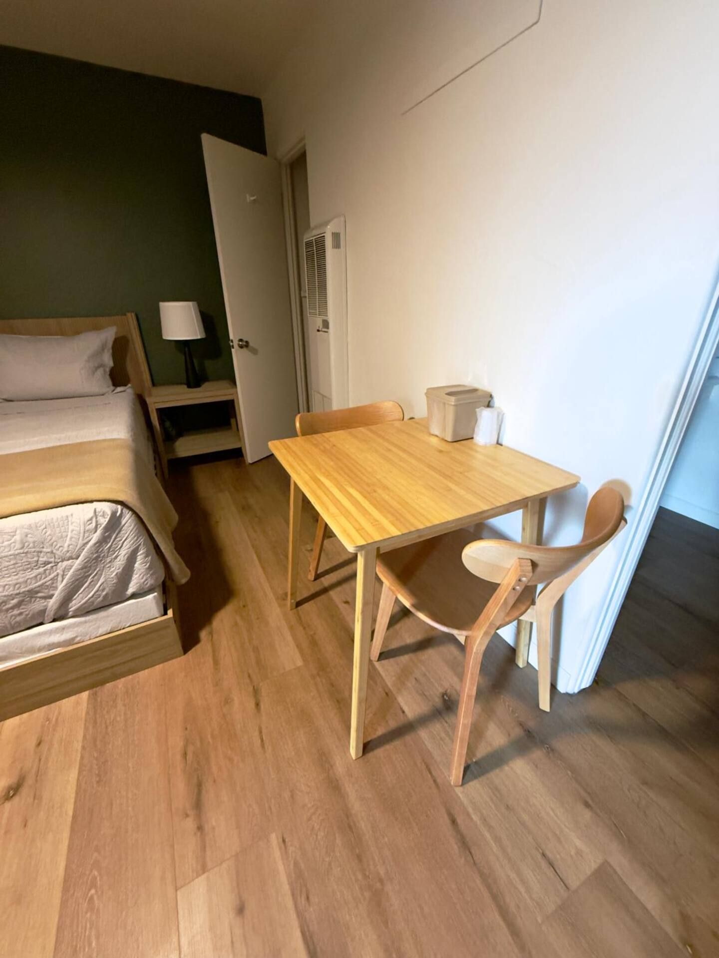 Um quarto pequeno e acolhedor, com uma cama, uma mesa de madeira, duas cadeiras, uma mesinha de cabeceira com abajur e uma porta.