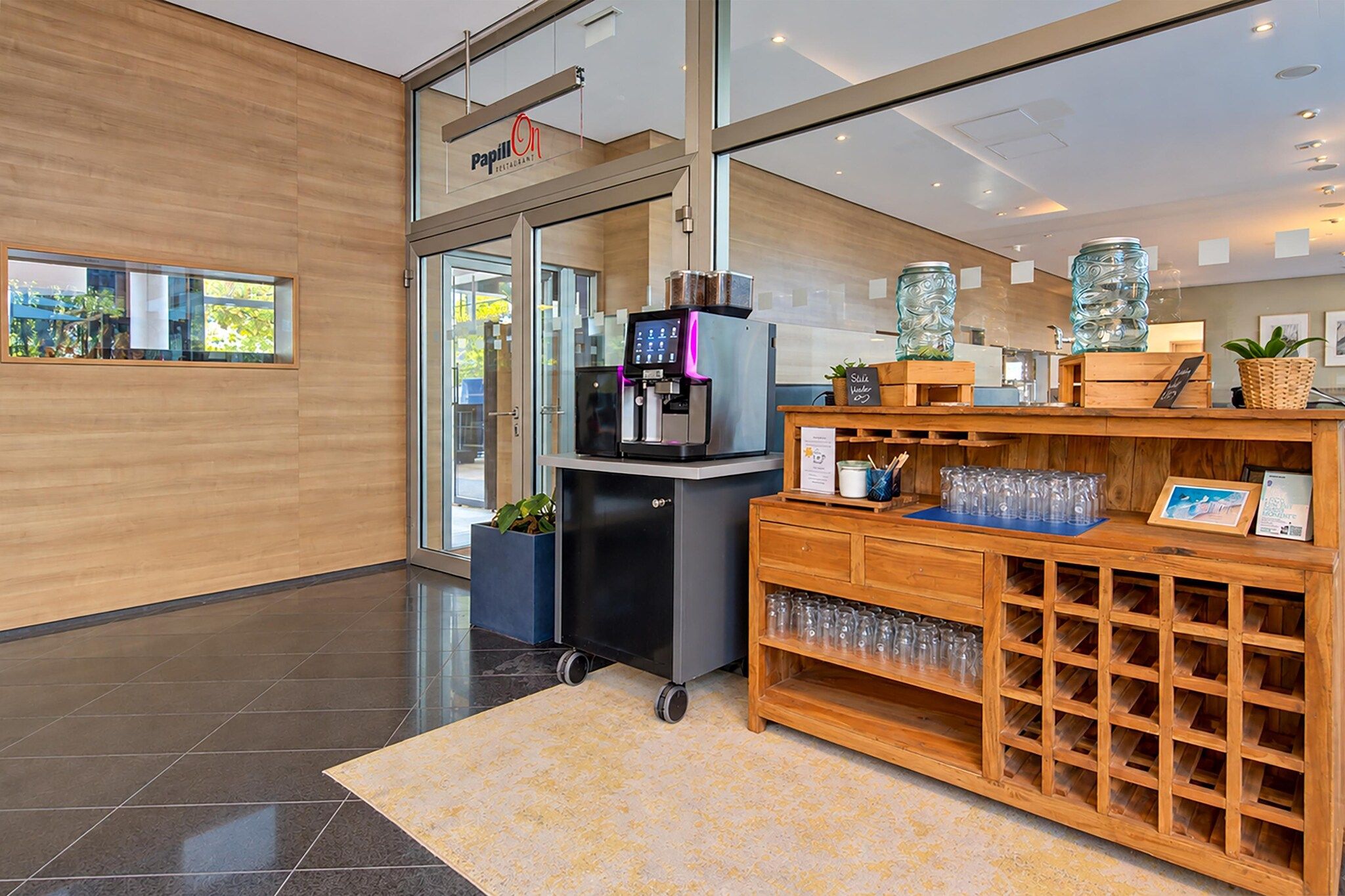 Un espace d’accueil moderne comprenant une machine à café, une vitrine et un comptoir en bois avec un porte-bouteilles à vin.