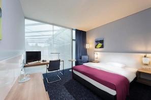 Habitación de hotel con cama, escritorio con televisor, silla, mesita y ventana con cortinas.