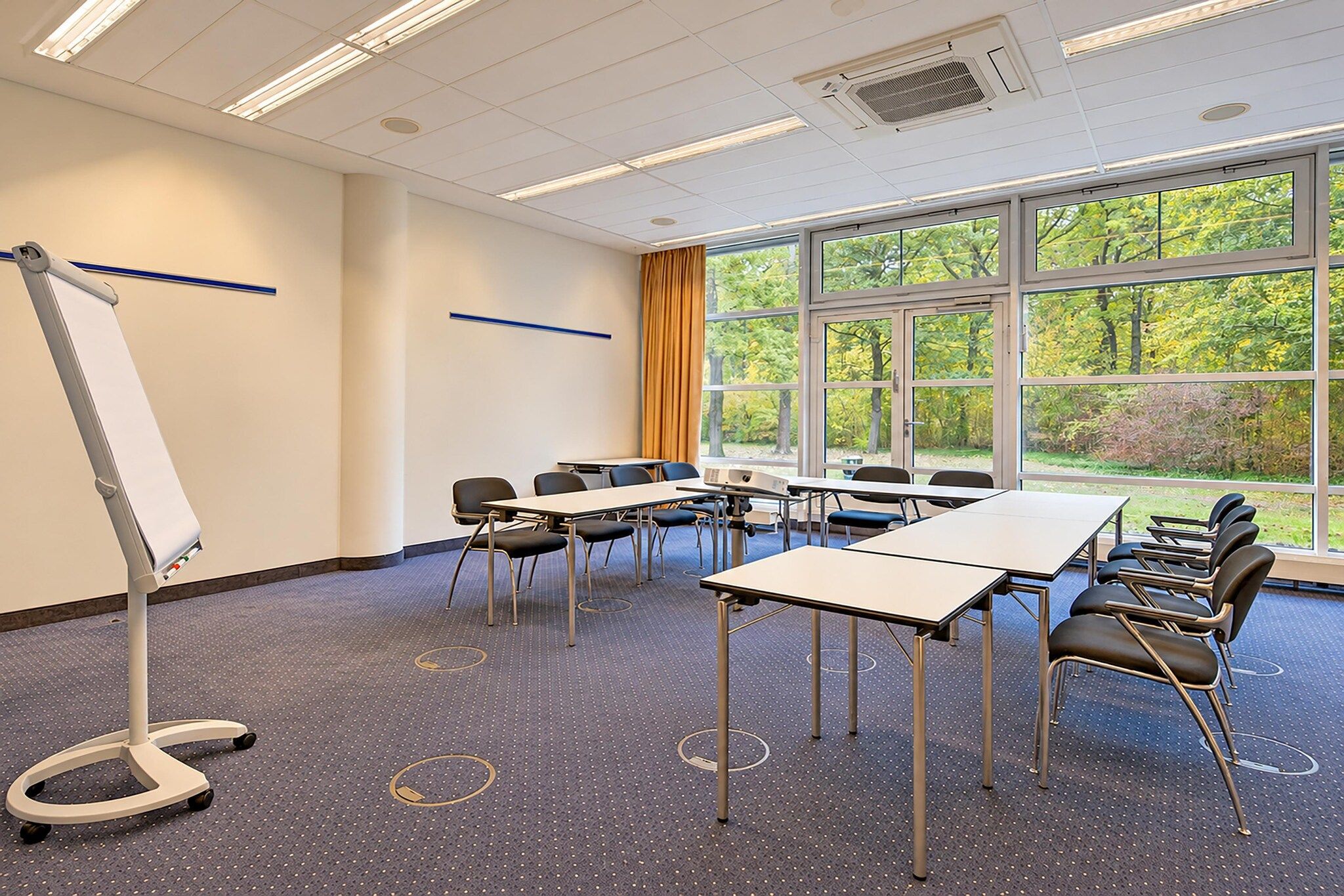 Ein Konferenzraum mit einer Wandtafel, Tischen, Stühlen und großen Fenstern mit Blick auf Bäume.