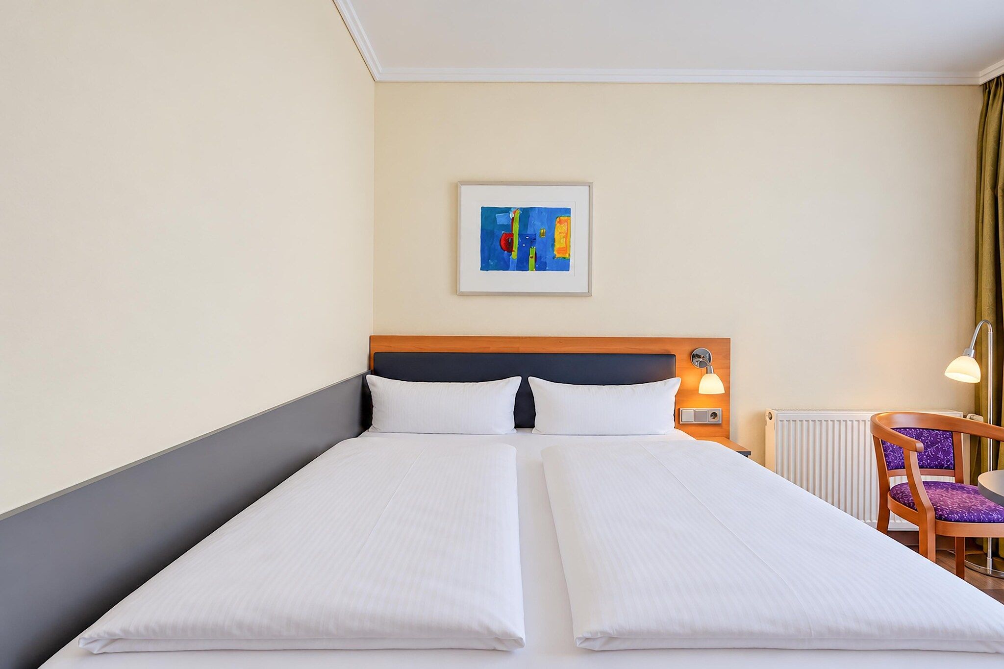 Ein Doppelbett mit weißen Laken, ein Kopfteil, ein Nachttisch mit Lampe, ein Sessel mit violettem Kissen und ein gerahmtes abstraktes Gemälde an der Wand.