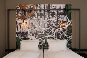 Una cama con ropa blanca y dos almohadas, un cabecero con un diseño abstracto complejo, y una pared con una mezcla de patrones y texturas.