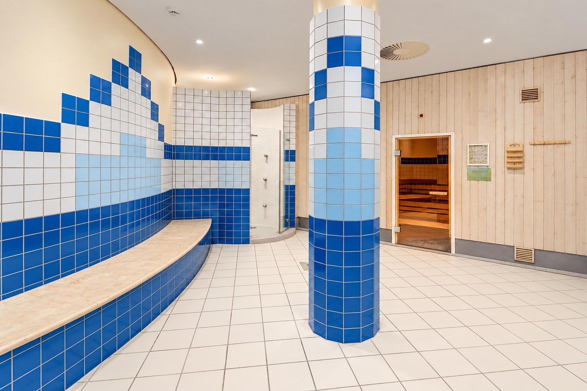 Ein modernes Badezimmer mit blauen und weißen Fliesen, einer Sitzbank und einer Holztür, die in einen anderen Raum führt.