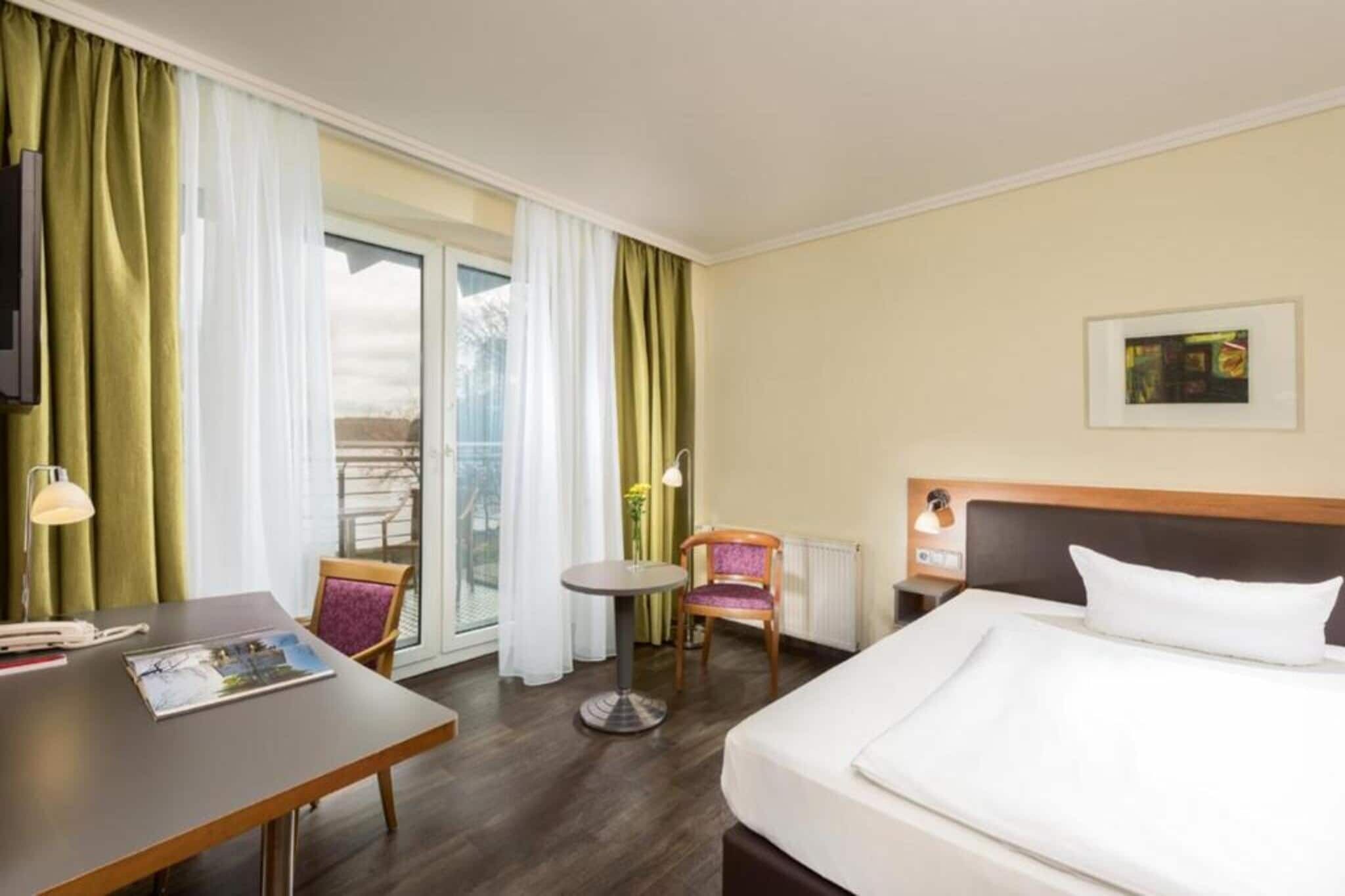 Una habitación de hotel con cama, escritorio, silla, una mesita y ventana con cortinas.