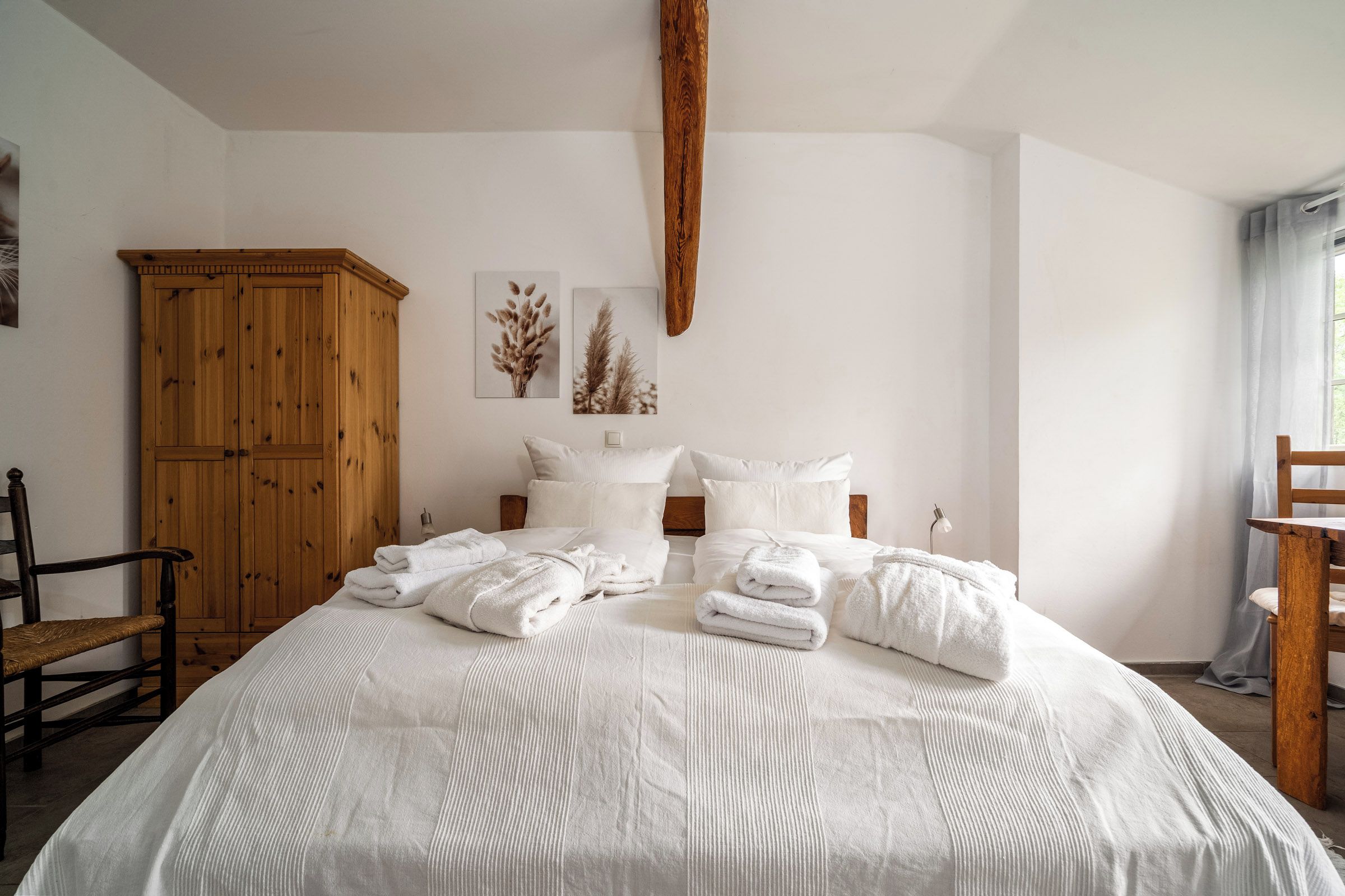 A bedroom with a large bed, a wooden wardrobe, a chair, and two wall-mounted pictures.