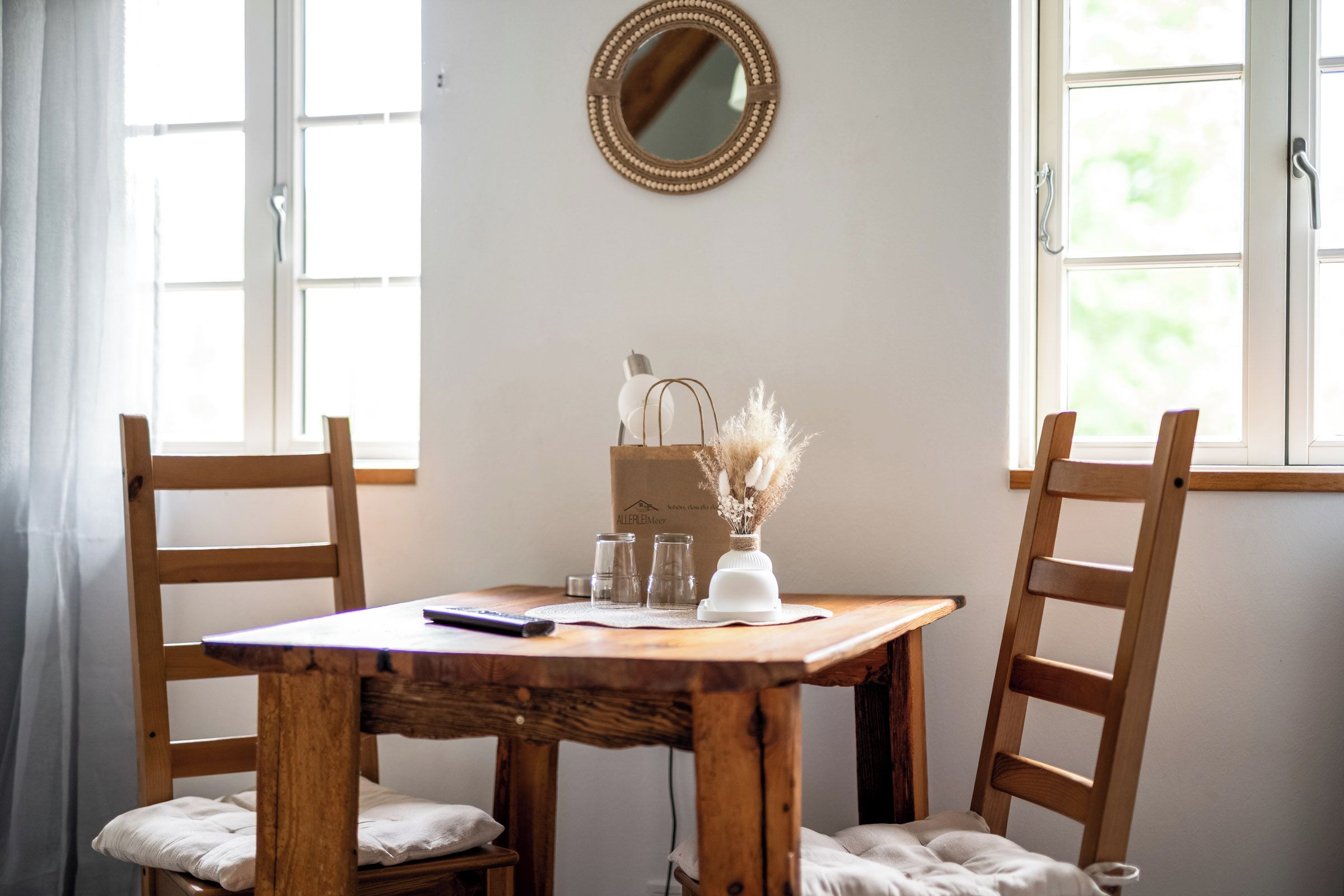 A wooden table with chairs, a mirror, and a vase with dried flowers.