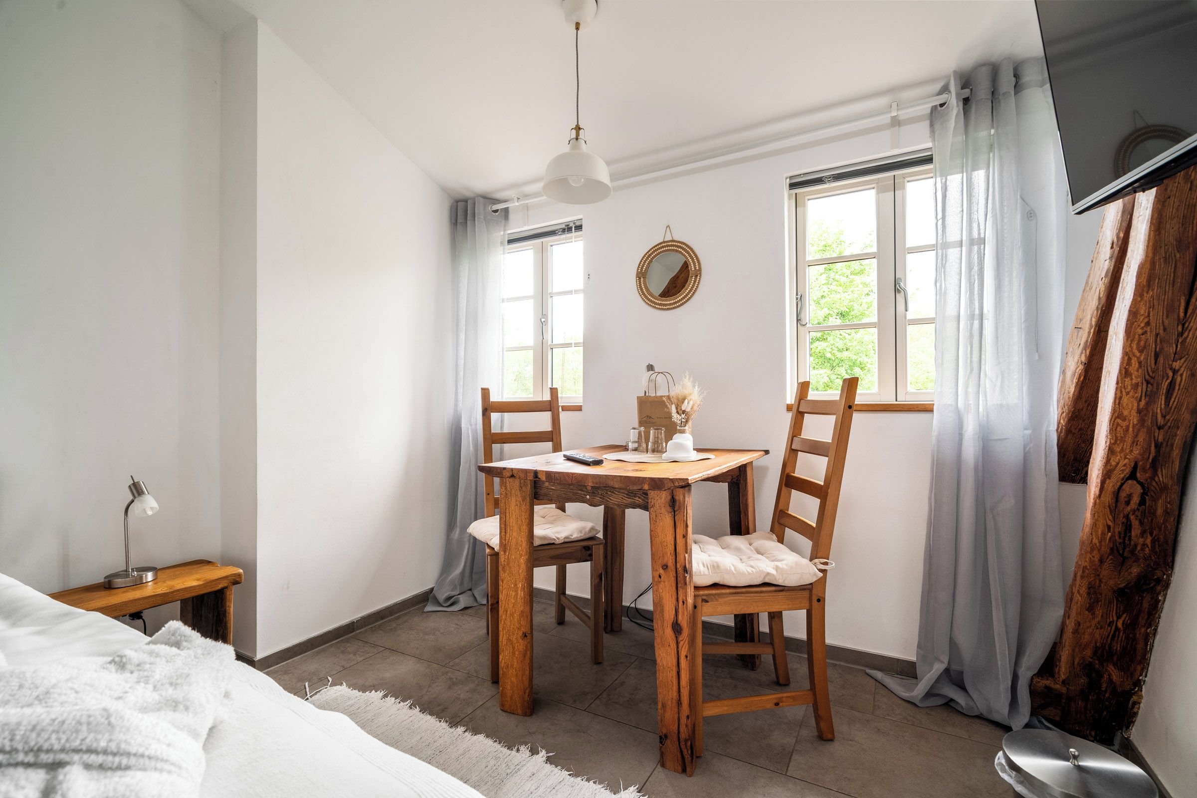 Une pièce avec une table en bois, deux chaises, un luminaire suspendu, un miroir rond et une fenêtre avec des rideaux.