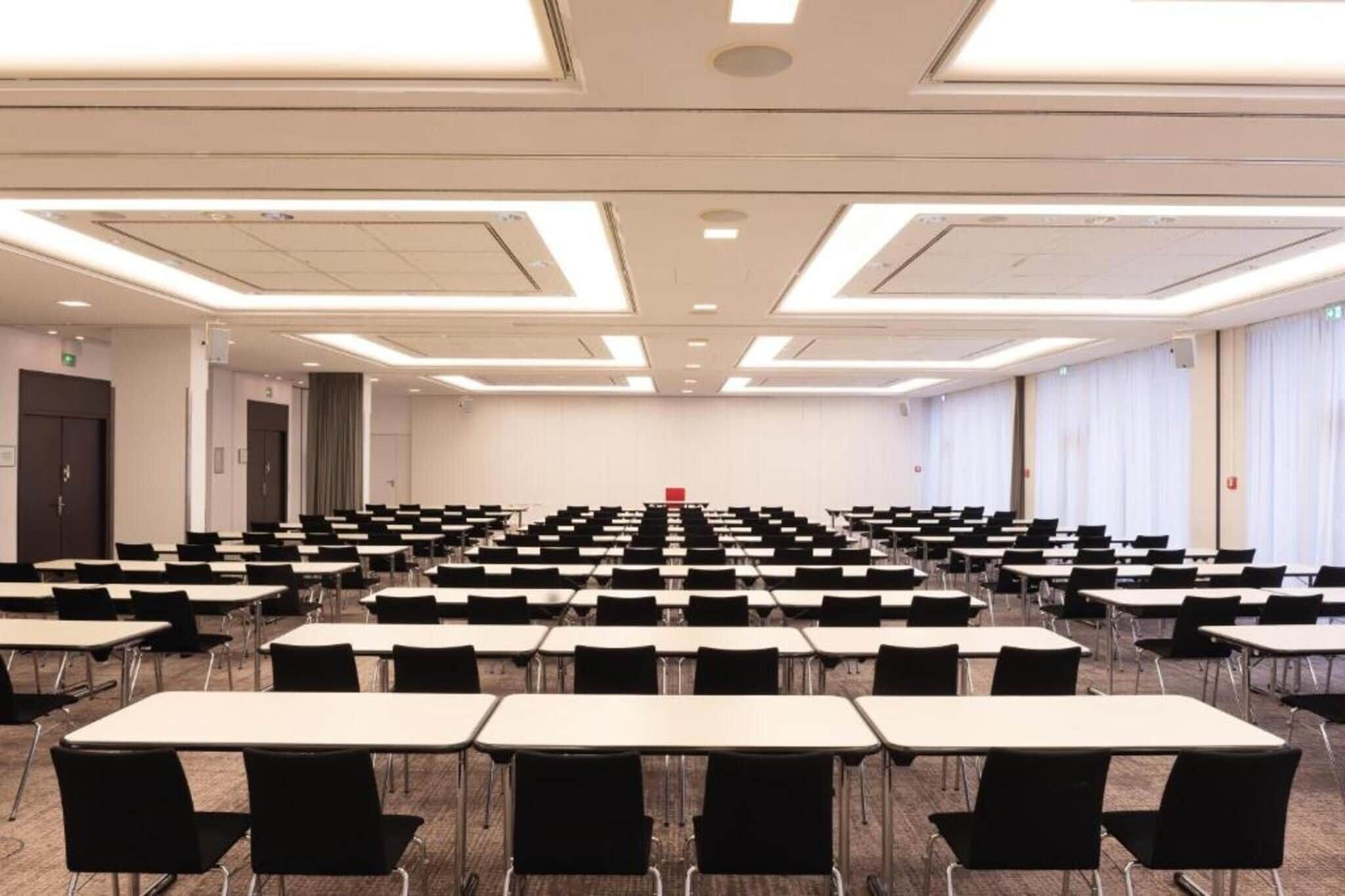 Un salón de conferencias con filas de mesas y sillas, un atril al frente y un ventanal grande en un lateral.