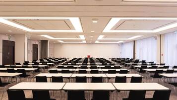 Un salón de conferencias con filas de mesas y sillas, un atril al frente y un ventanal grande en un lateral.