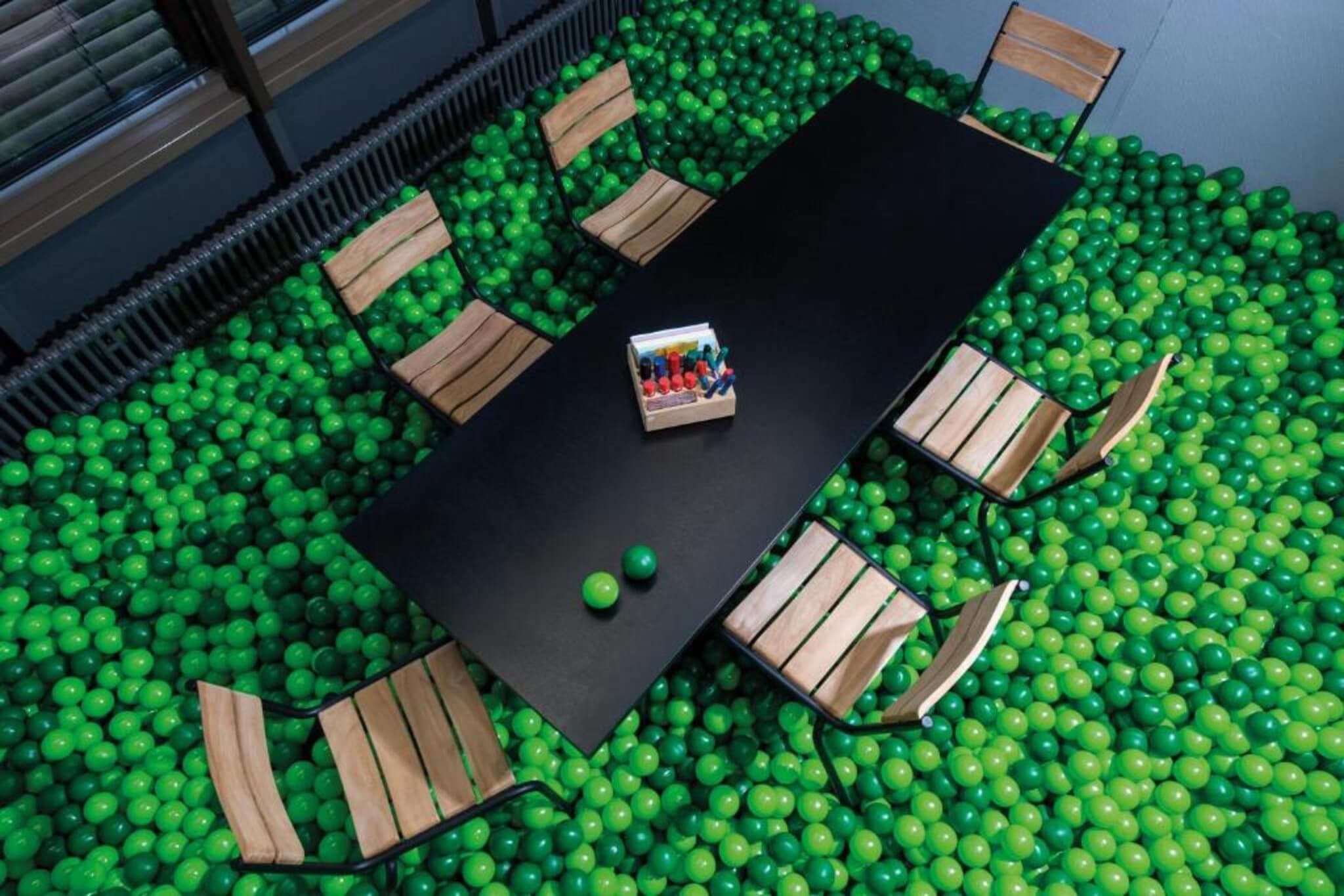 A room with a black table surrounded by wooden chairs, set in a ball pit filled with green balls.