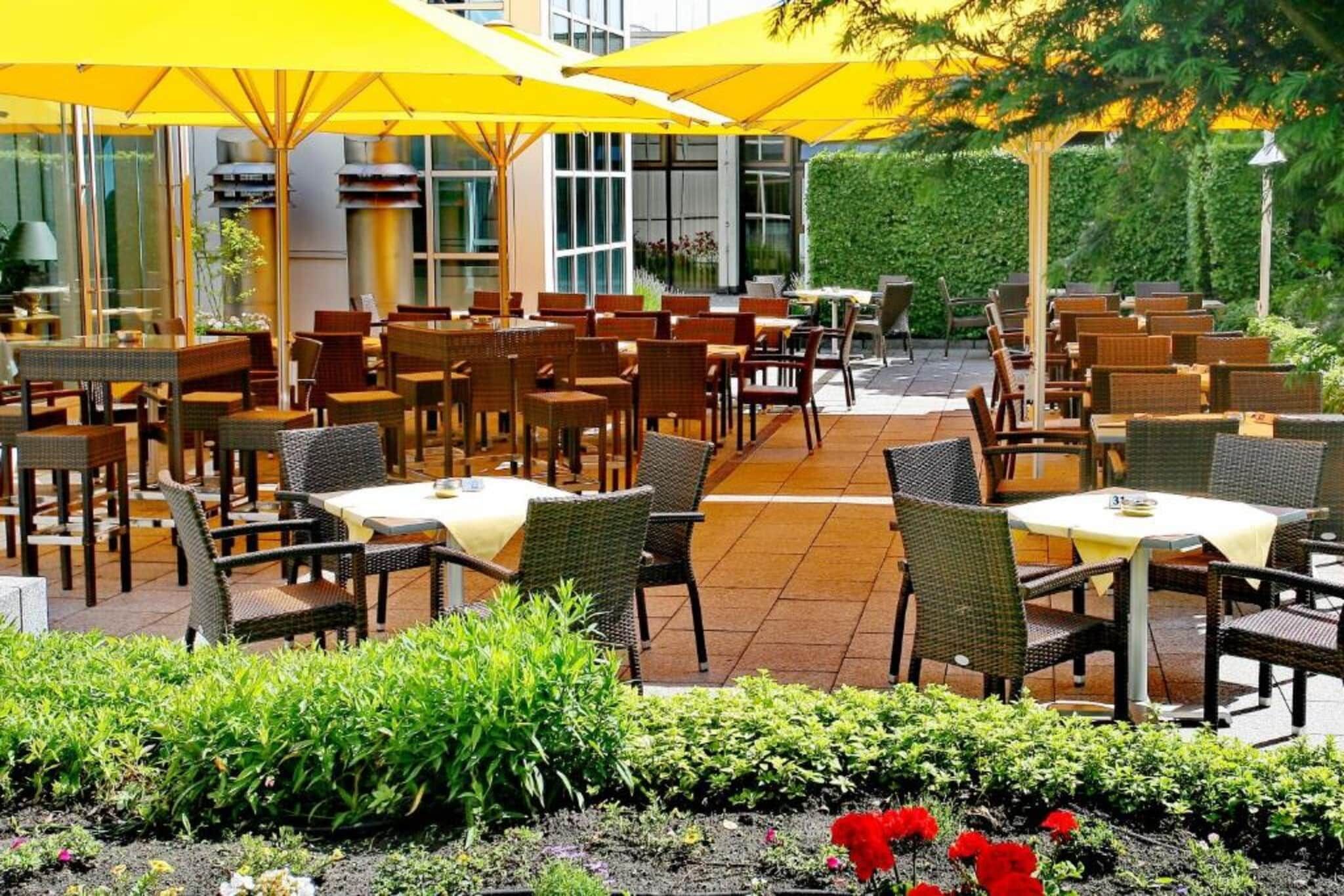 Ein Außenbereich zum Essen mit Tischen und Stühlen, gelben Sonnenschirmen und einem Garten mit Blumen.