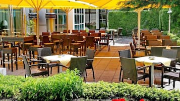 Ein Außenbereich zum Essen mit Tischen und Stühlen, gelben Sonnenschirmen und einem Garten mit Blumen.