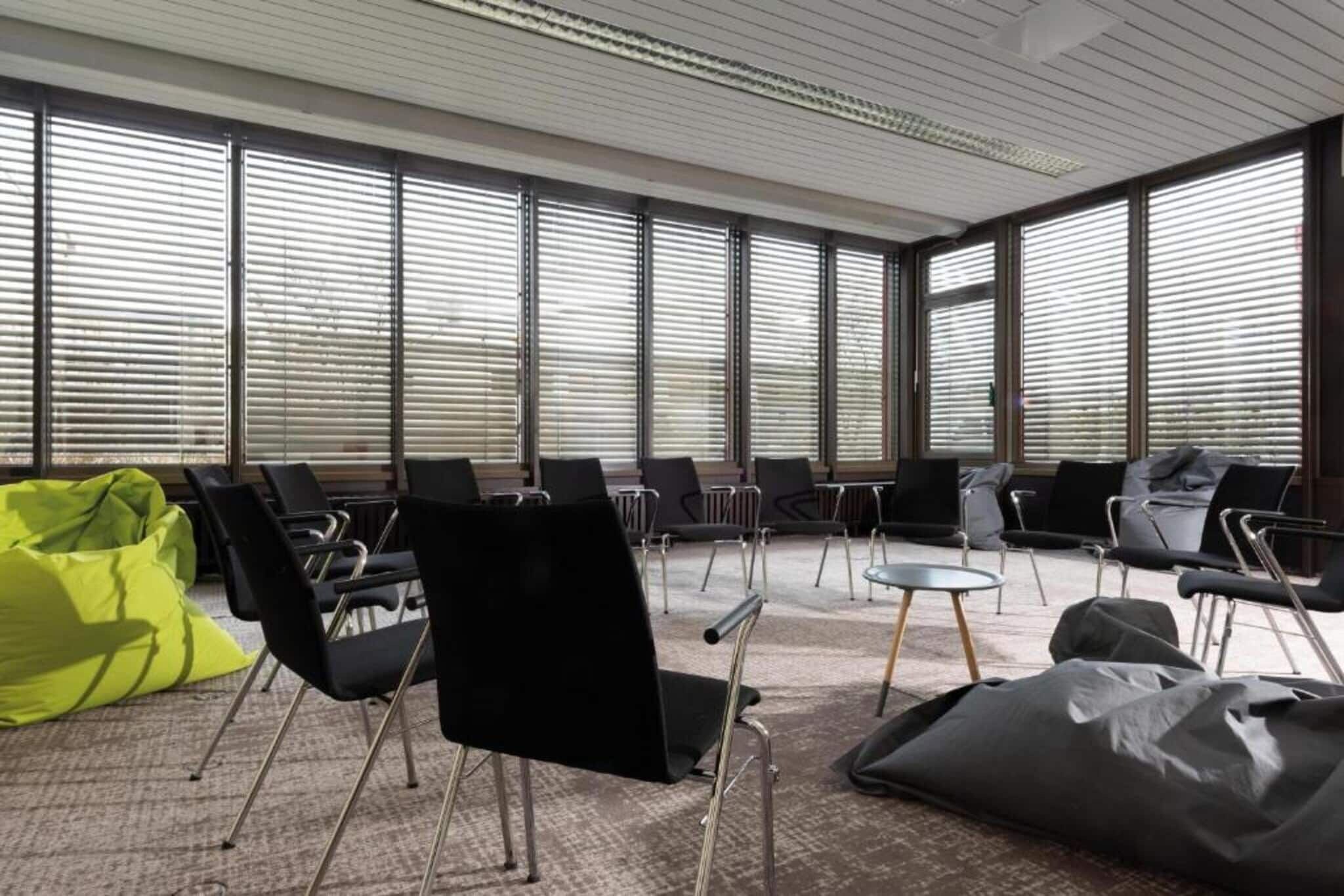 Een vergaderzaal met vloerbedekking, meerdere zwarte stoelen, een ronde tafel en grote ramen met jaloezieën.