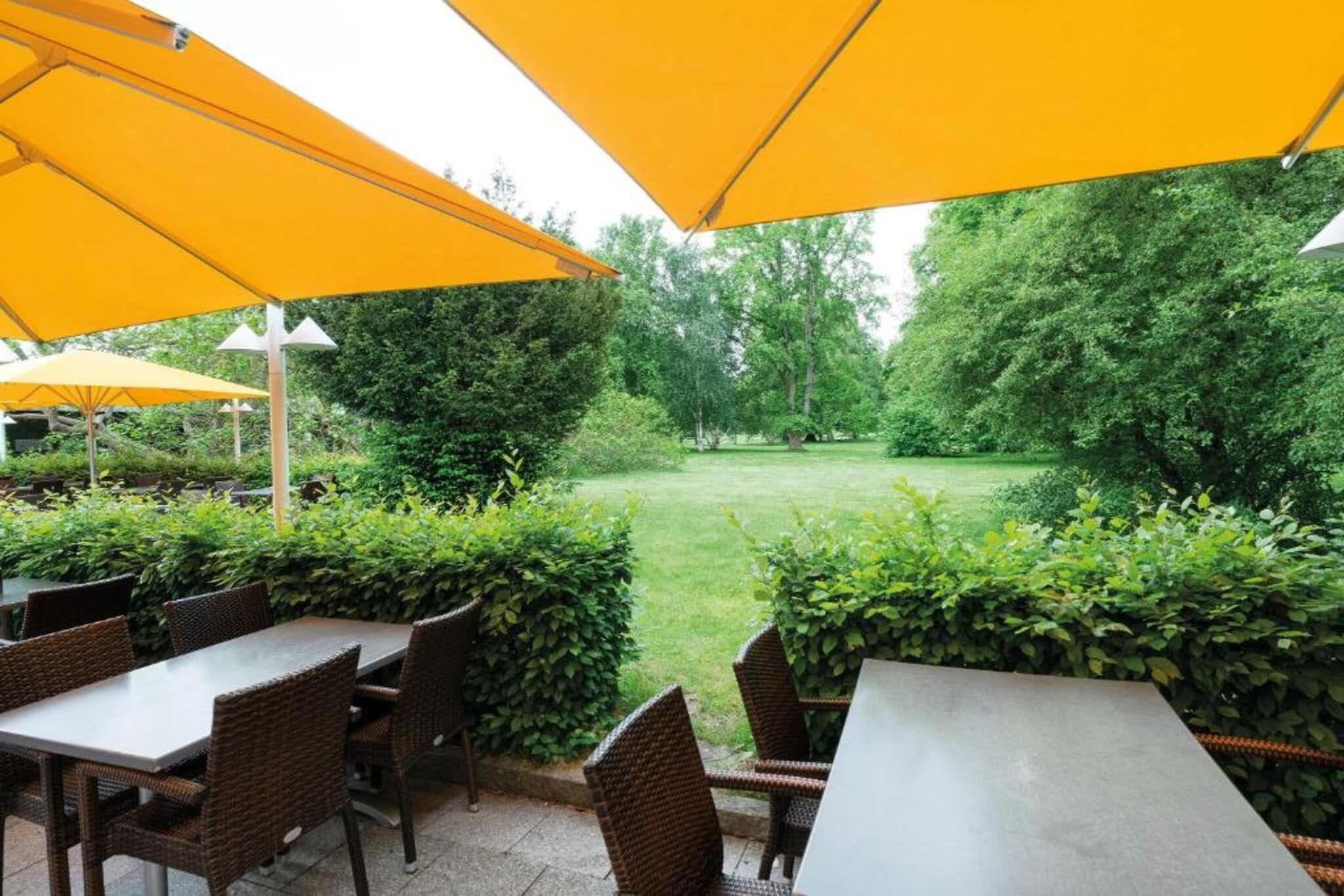 An outdoor seating area with tables and chairs under yellow umbrellas, surrounded by greenery and trees.