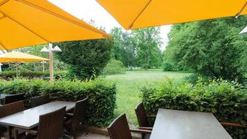 An outdoor seating area with tables and chairs under yellow umbrellas, surrounded by greenery and trees.
