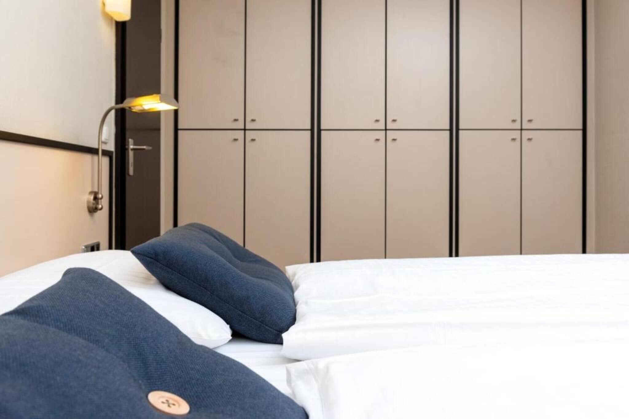 Una cama con ropa de cama blanca y azul, una lámpara fijada a la pared y un armario con varias puertas.