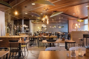 Un intérieur de restaurant moderne avec des panneaux de plafond en bois, des luminaires suspendus et différents types d’aménagements pour s’asseoir, notamment des tables et des chaises.
