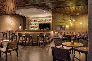 Zona de bar con diversas opciones de asientos, incluyendo taburetes y mesas, una barra con gran variedad de botellas y una estantería mural con botellas.