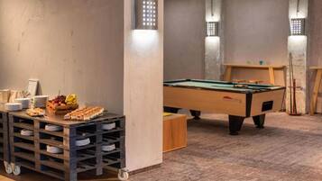 A room with a pool table, a snack bar with plates and fruit, and a billiards table.