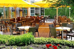 Un área de comedor al aire libre con mesas y sillas, sombrillas amarillas y un jardín con flores.
