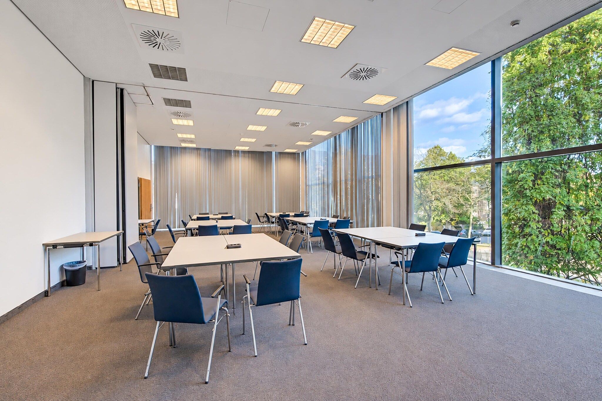 Une salle de conférence équipée de tables et de chaises, dotée de grandes fenêtres et offrant une vue sur les arbres.