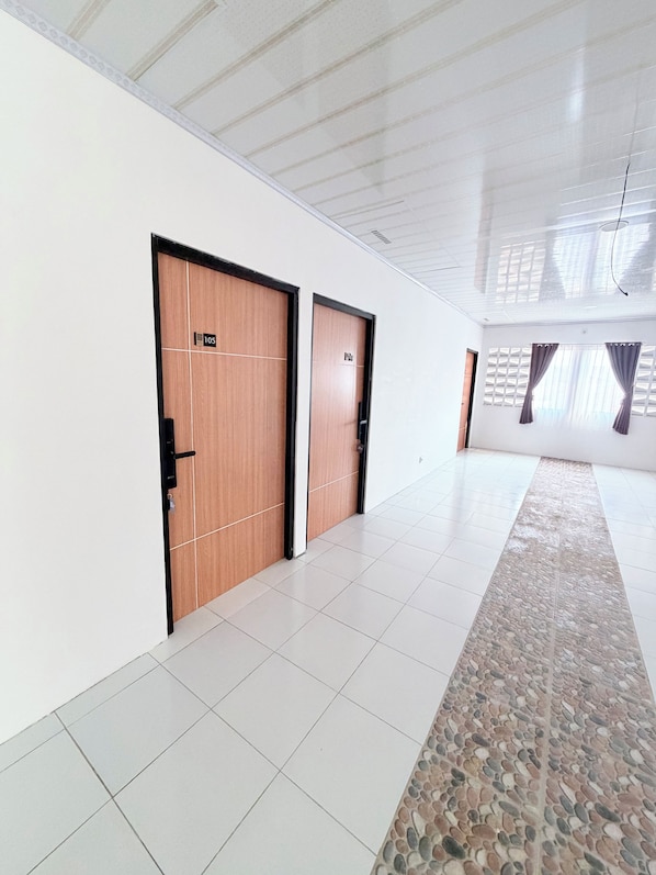 Un couloir avec deux portes en bois, chacune portant un numéro. Le sol est carrelé, et un tapis à motifs mène à une fenêtre ornée de rideaux.