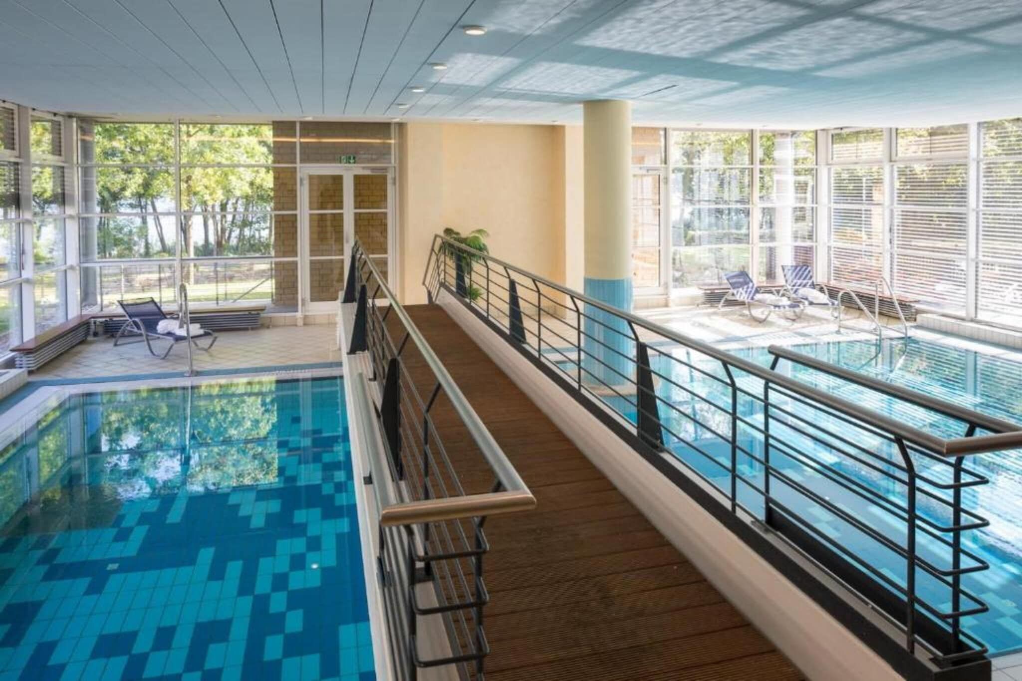 Ein Hallenbad mit Holzterrasse, Glasgeländern und Liegestühlen.